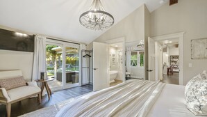 A spacious bedroom with a large bed, a chandelier, and a view of the garden through the sliding glass doors.