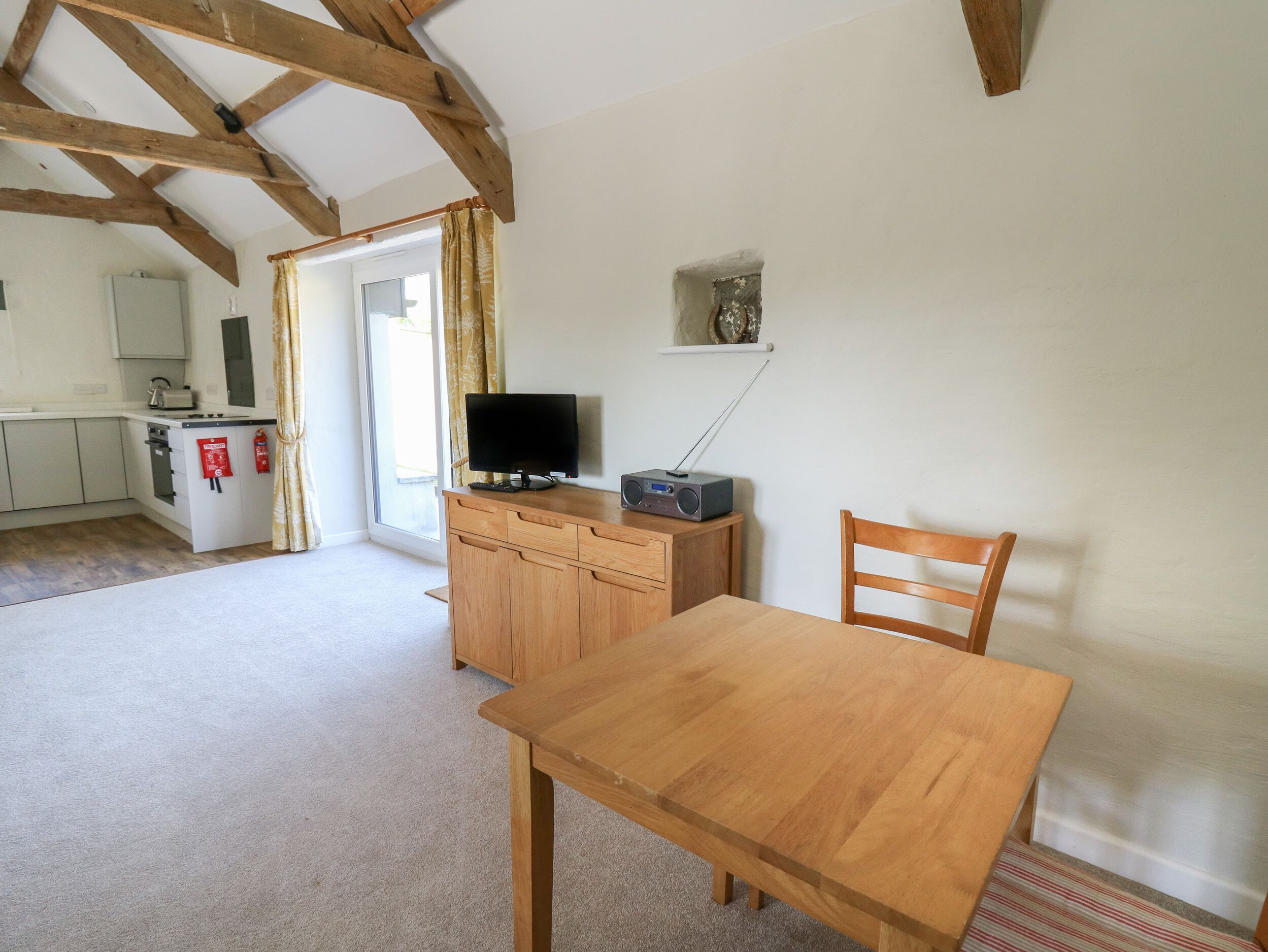 A room with a wooden table and chair, a television, and a kitchen in the background.