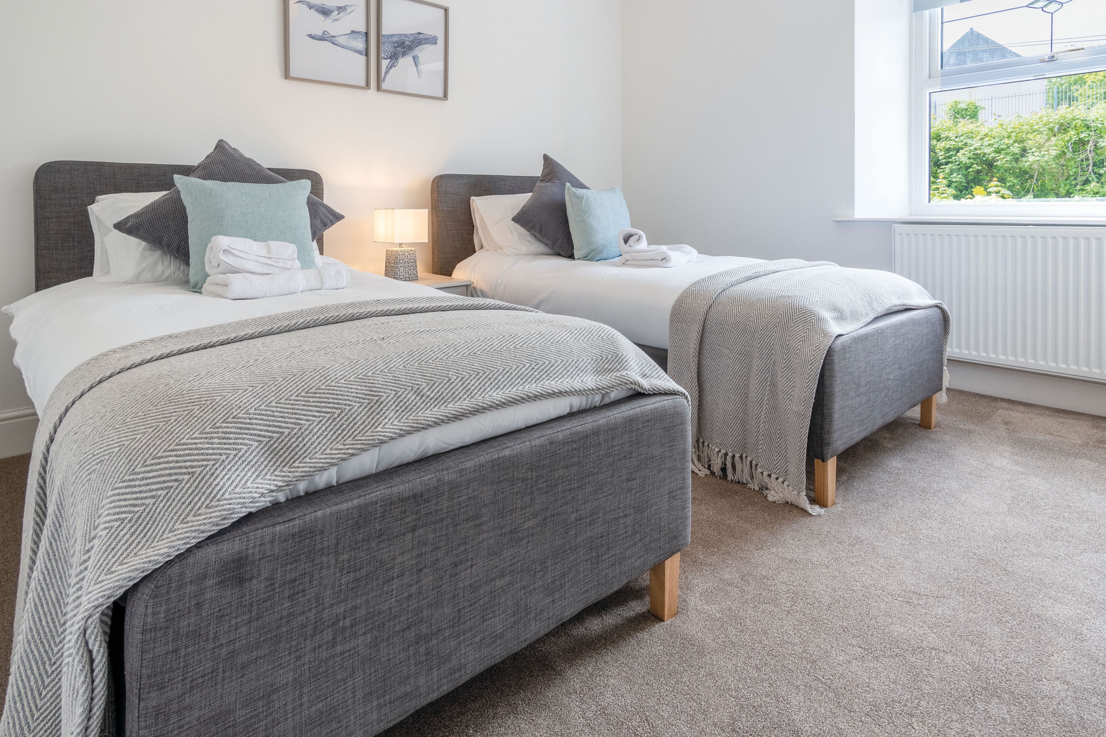 A bedroom with two beds, each with a grey headboard, white bedding, and a grey throw blanket. There is a window with a view of greenery outside.