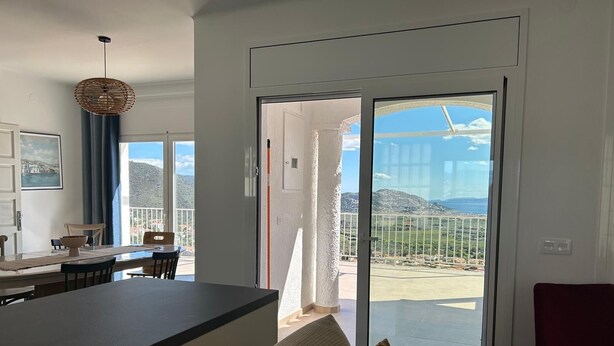 A modern kitchen with a dining area, a large sliding glass door leading to a balcony, and a view of the surrounding landscape.