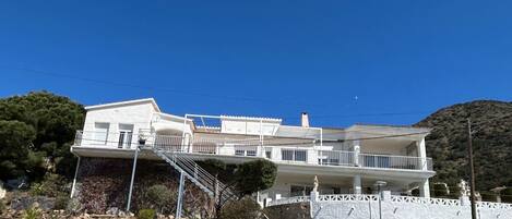 A multi-story white building with balconies, a staircase, and a view of a mountainous landscape.