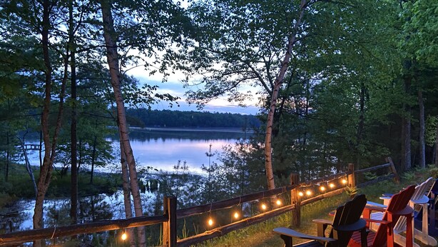Un área de descanso junto al lago con sillas y una mesa de madera, rodeada por una valla de madera y luces colgantes.