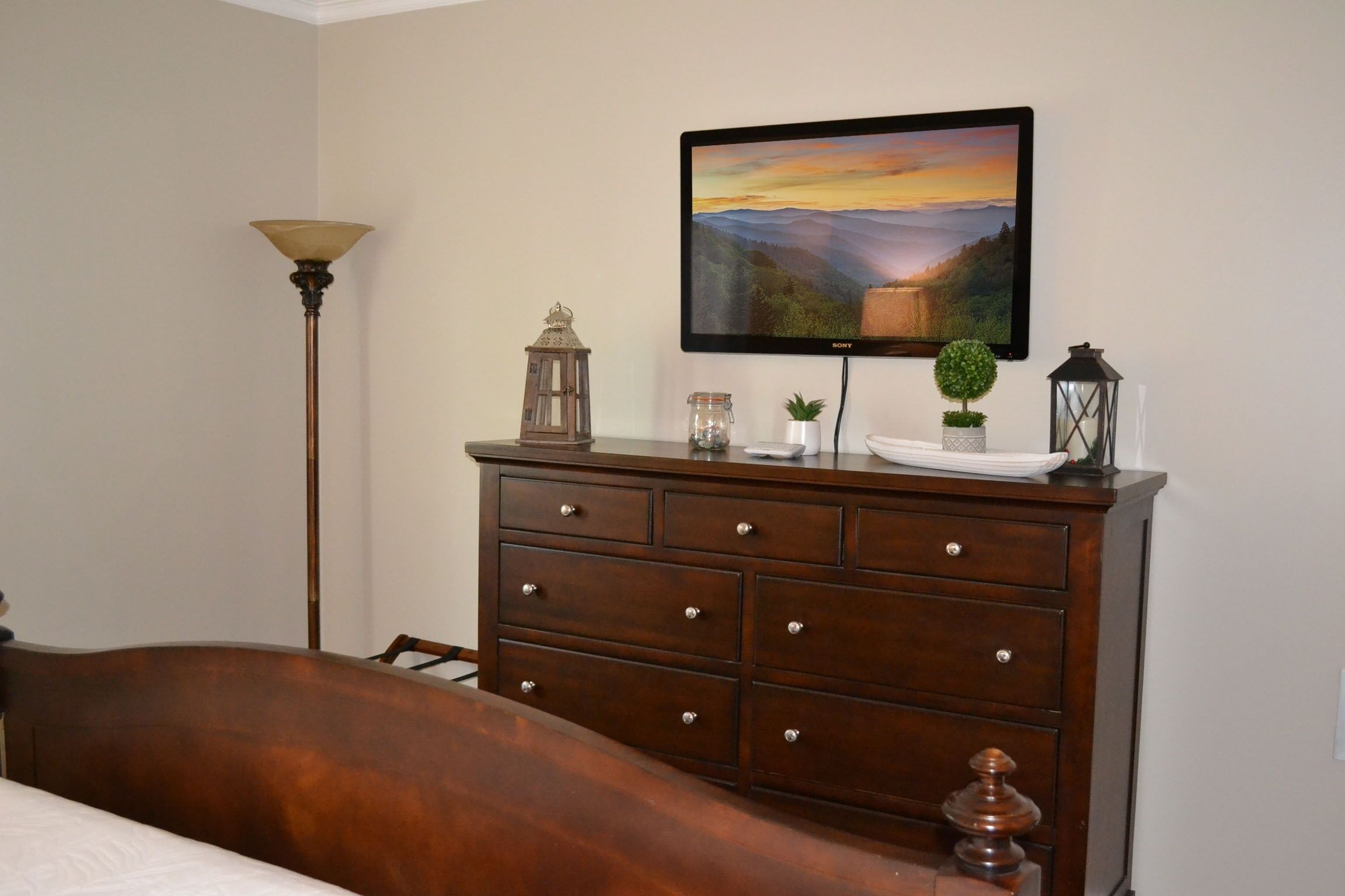 A bedroom with a wooden dresser, a bed, a lamp, a TV mounted on the wall, and a landscape painting.