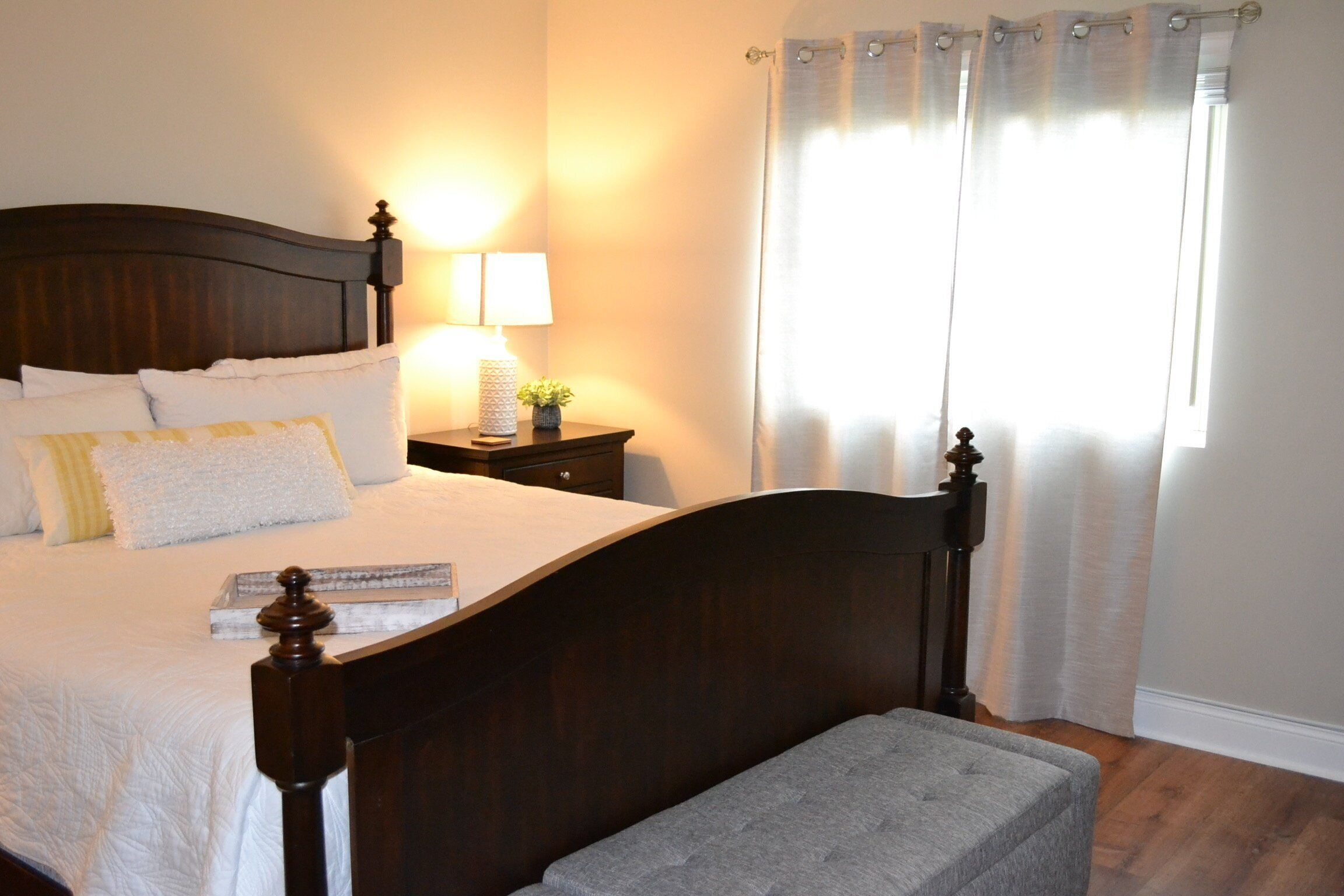 A neatly made bed with a wooden headboard, a bedside table with a lamp, and a window with curtains.
