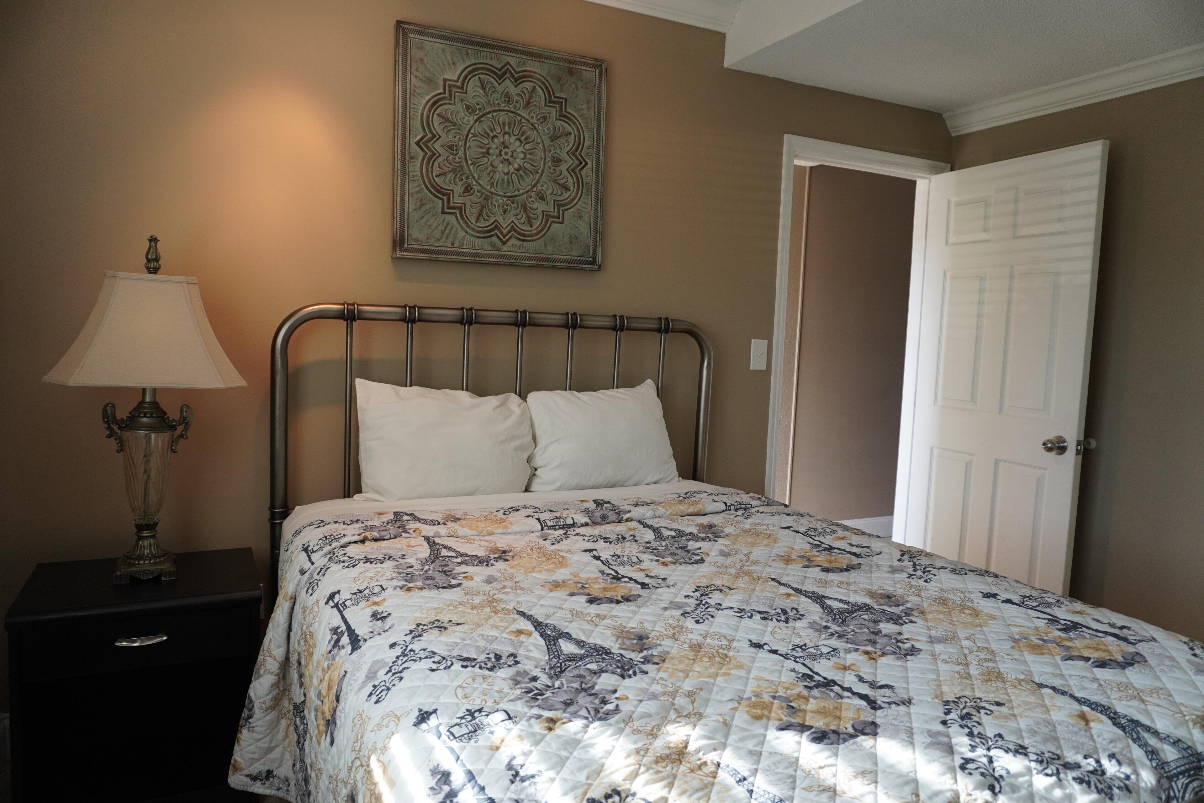 A bedroom with a bed, a nightstand, a lamp, a door, and a decorative wall piece.