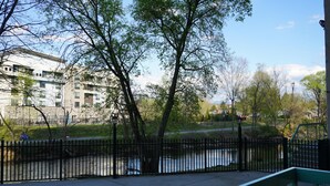 Een voetpad langs de rivier met een metalen hek, bomen en een gebouw op de achtergrond.