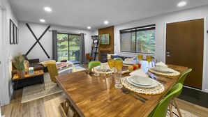 Uma sala de jantar com mesa de madeira posta para quatro pessoas, cadeiras verdes e vista para o exterior através de grandes janelas.