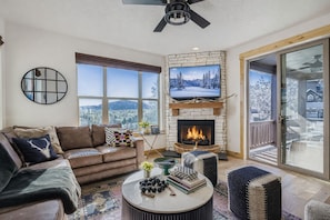 A cozy living room with a fireplace, a comfortable sofa, a coffee table with books and decorative items, and a large window offering a scenic view.