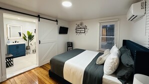 Ein Schlafzimmer mit Bett, Fernseher, einer Schiebetür zum Badezimmer und einem Fenster mit Jalousien.