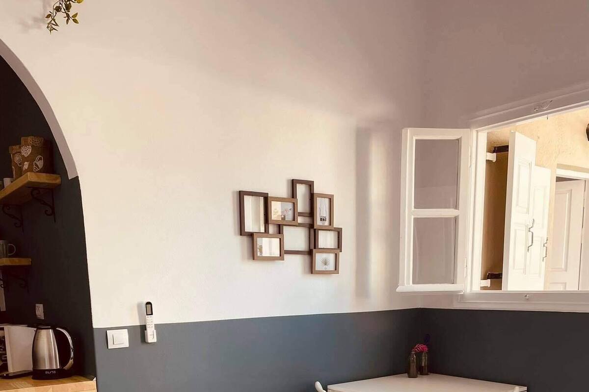 A bedroom with a bed, a small table, and a chair. There is a window with a view of a door and a plant.