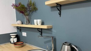 Eine Küche mit Spüle aus Edelstahl, Wasserkocher und Kaffeemaschine. Auf einem Regal stehen zwei weiße Tassen und ein Blumenvasen.