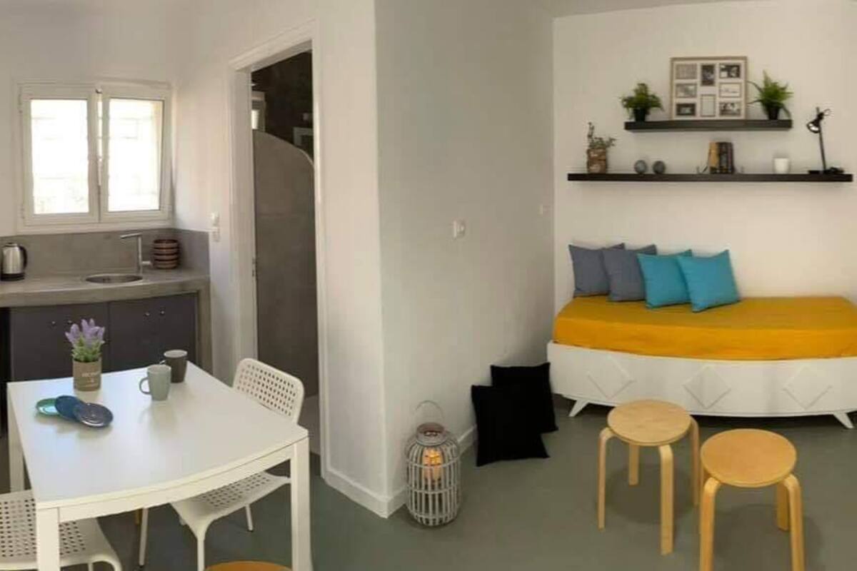 A compact living space with a kitchenette, a bed with a yellow headboard, and a small dining area.