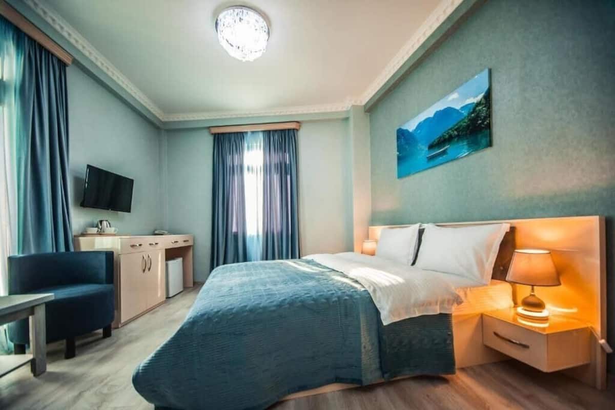 A hotel room with a bed, bedside table, lamp, TV, desk, chair, and curtains.