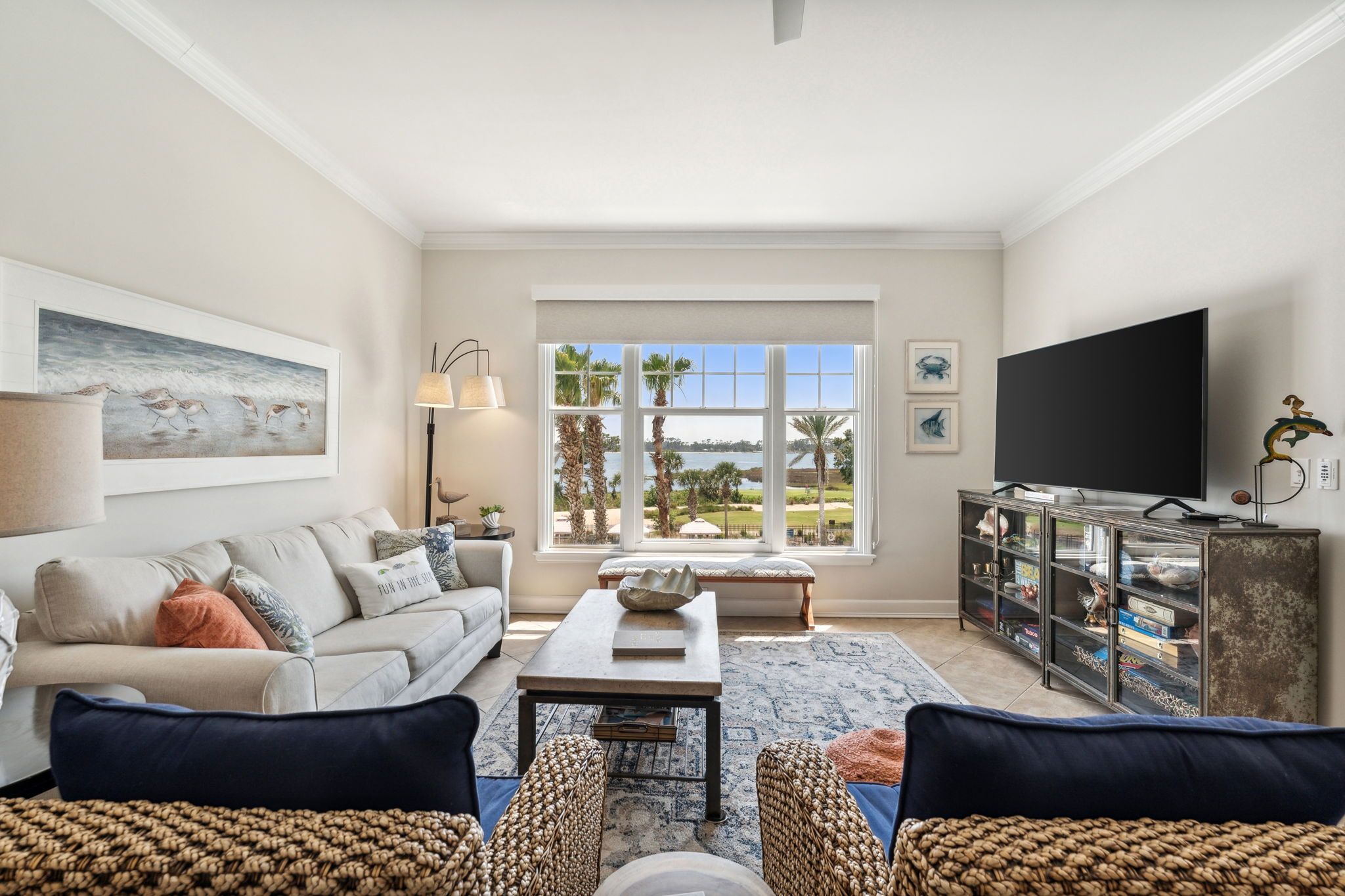 A living room with a sofa, a coffee table, a TV, and a view of palm trees through large windows.
