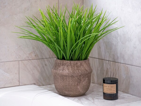 Una planta en maceta con hojas verdes largas en una maceta texturizada, junto a una vela sobre una superficie con baldosas.