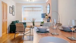 Una sala de estar moderna con una mesa de comedor preparada para dos personas, un sofá verde, una silla de mimbre y una mesita de noche con una lámpara.
