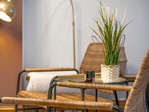 Una silla de mimbre con asiento y respaldo trenzados, una pequeña mesa redonda con base trenzada y una planta en maceta con hojas verdes largas y flores blancas.