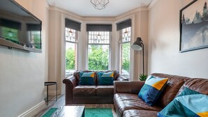 A living room with a brown leather sofa, a wooden coffee table, and a green rug. There are two windows with decorative glass panes and a painting on the wall. - Beautiful 4BR Restored Red Brick Cafes and  Park - Pass the Keys (Belfast)
