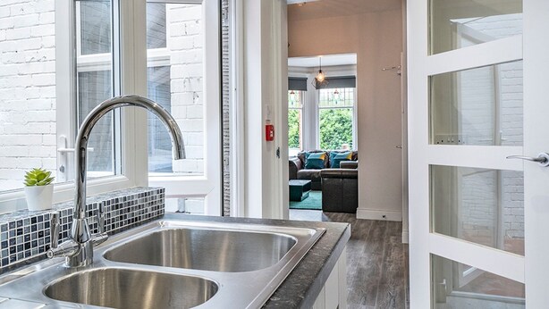 A kitchen with a double sink, a potted plant, and a view to a living area with a sofa and a hanging light fixture. - Beautiful 4BR Restored Red Brick Cafes and  Park - Pass the Keys (Belfast)