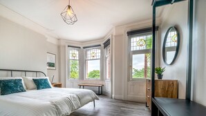 A bedroom with a large bed, a wooden floor, a desk, and a dresser. There are windows with decorative glass panes and a view of greenery outside. - Beautiful 4BR Restored Red Brick Cafes and  Park - Pass the Keys (Belfast)