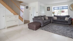 A spacious living room with a grey sofa set, a matching ottoman, and a large window providing natural light. A staircase with wooden railings leads to an upper level.