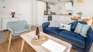 Una sala de estar moderna con un sofá azul, una mesita con una botella y vasos, una silla de mimbre y una cocina al fondo.