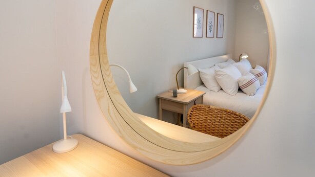 Un dormitorio con un espejo circular, una cama con ropa de cama blanca, una mesita de noche con lámpara y una silla de mimbre.