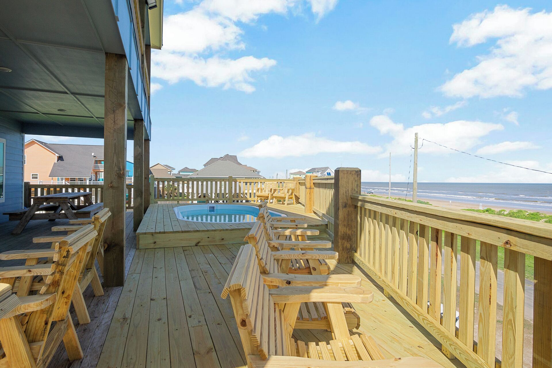 A wooden deck with a hot tub, wooden chairs, and a view of the beach.