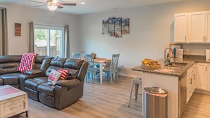 A modern living room with a leather sectional sofa, dining area, kitchen with white cabinets, and a ceiling fan. - Clean New Townhome: Comfort and Convenience Await! (Hampstead)