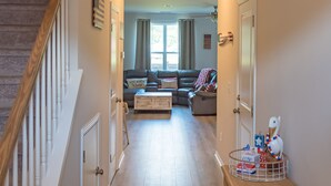 A hallway with a wooden staircase, a wooden table with books and a telephone, and a living area with a sofa and a window. - Clean New Townhome: Comfort and Convenience Await! (Hampstead)