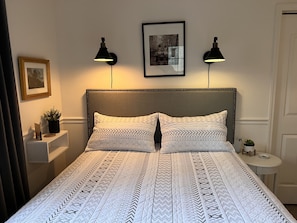 A neatly made bed with patterned bedding, two wall-mounted reading lamps, a framed photograph on the wall, and a small bedside table with a plant.