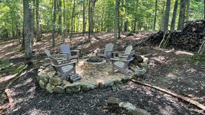 A forested area with a fire pit surrounded by Adirondack chairs, a stone fire ring, and a stack of firewood.