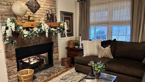 A cozy living room with a brick fireplace, a brown sofa, a glass coffee table, and a large window with curtains.