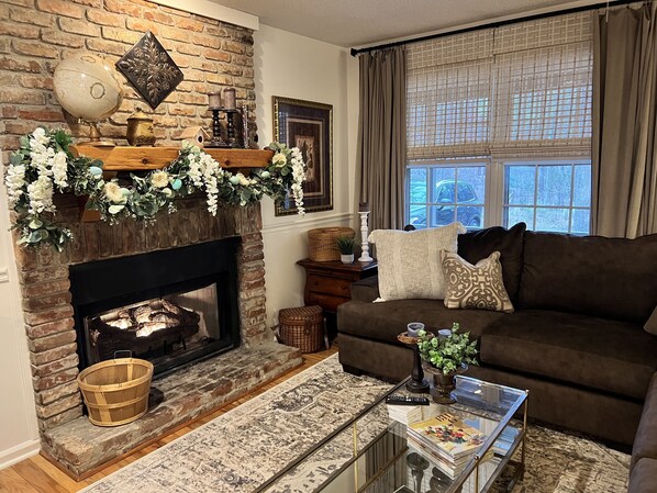A cozy living room with a brick fireplace, a brown sofa, a glass coffee table, and a large window with curtains.
