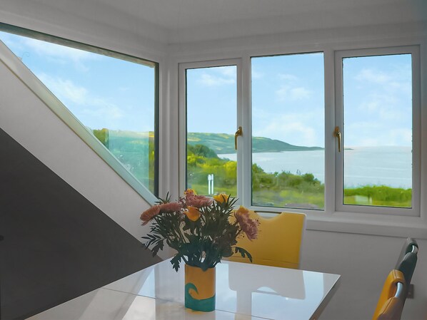 A dining area with a table, chairs, and a vase of flowers. There are large windows with a view of a body of water and greenery.
