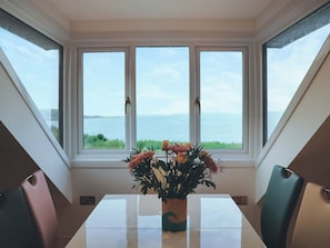 A dining table with a vase of flowers and a view of the sea through large windows.