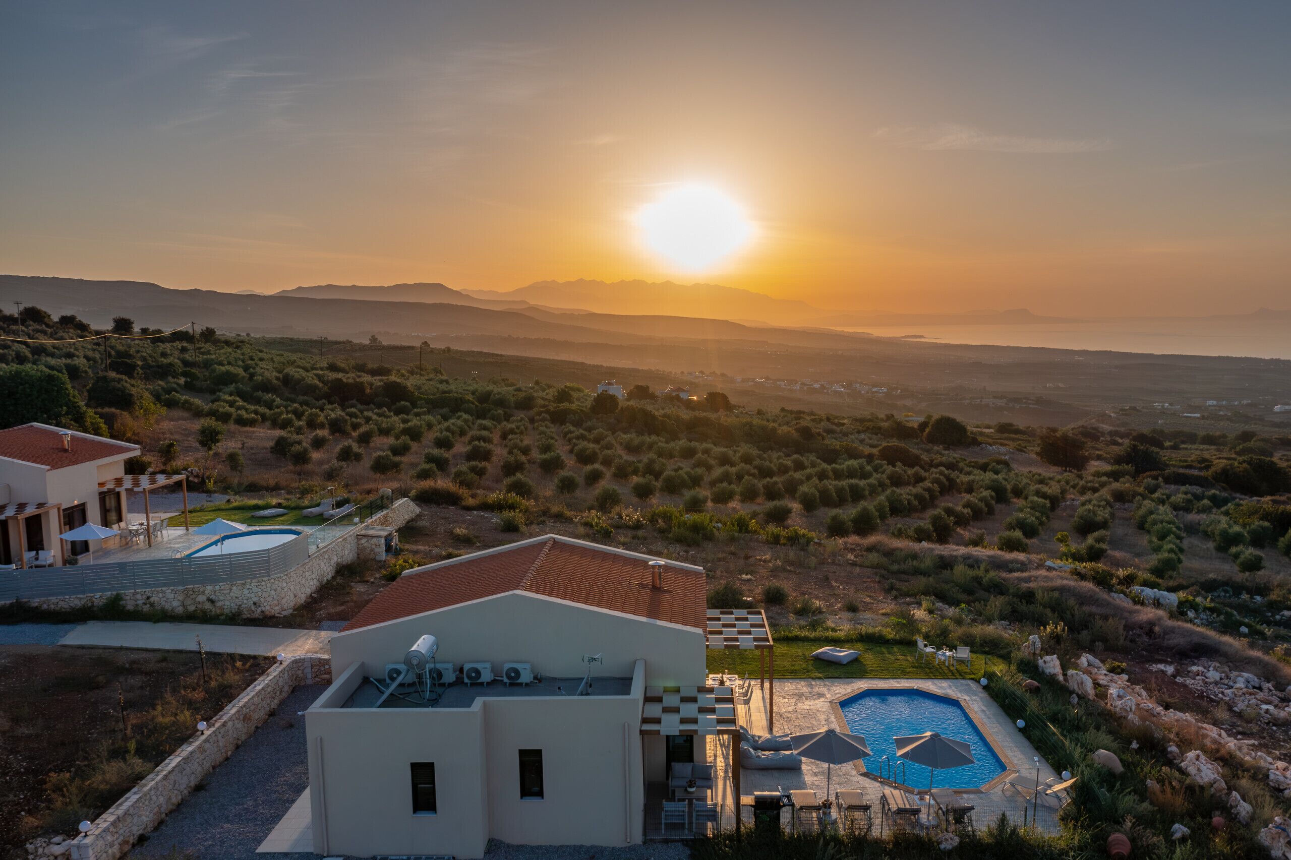 A modern villa with a swimming pool, outdoor seating, and a view of the surrounding landscape during sunset.