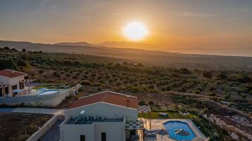 A modern villa with a swimming pool, outdoor seating, and a view of the surrounding landscape during sunset.