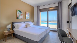 A hotel room with two beds, a desk, a chair, and a large window with a view of the beach. - Jason Panorama 1 Villa (Rethymno)