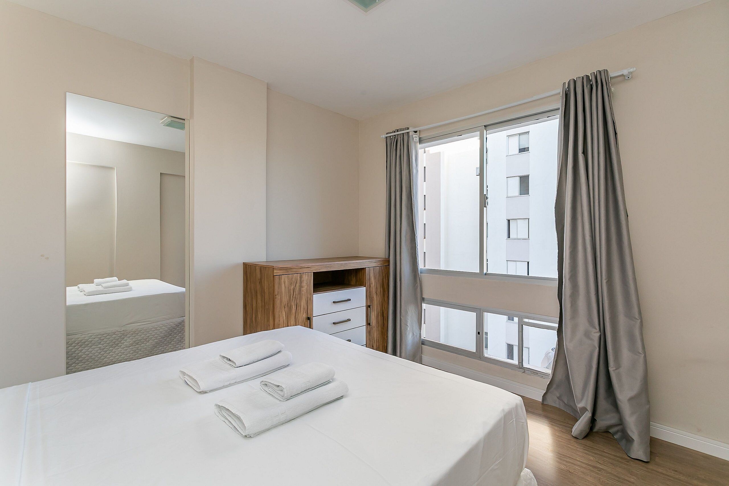 Une chambre d’hôtel avec un lit, un bureau en bois, un miroir et une fenêtre avec des rideaux.