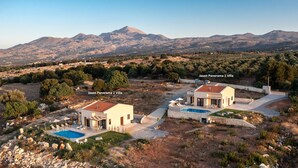 Two villas with swimming pools, surrounded by rocky terrain and olive trees, with mountains in the background.