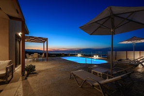 A rooftop pool area with lounge chairs, umbrellas, and a view of the ocean at dusk.