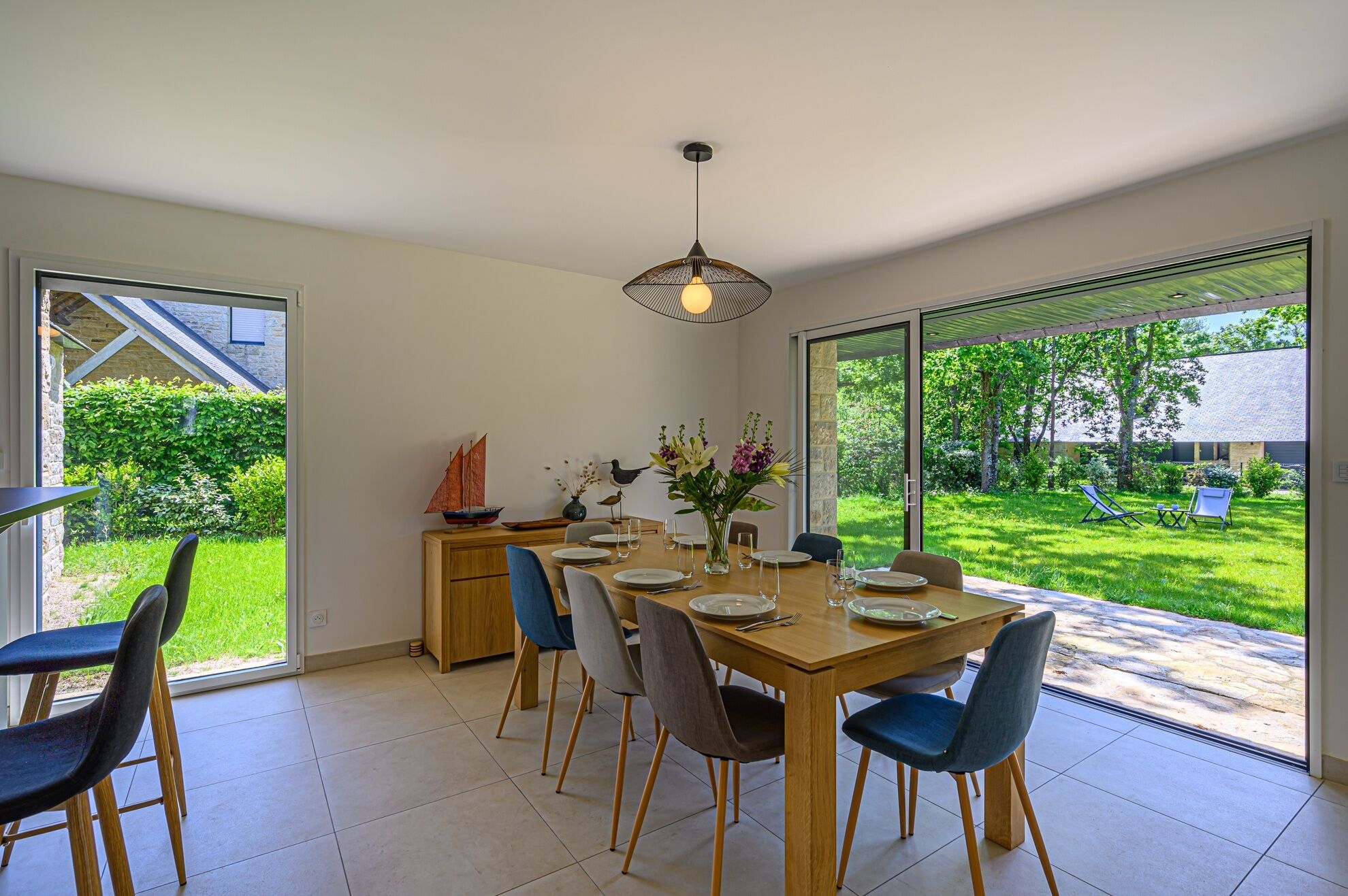 Un coin repas avec une table en bois dressée pour quatre personnes, entourée de chaises, et offrant une vue sur un jardin luxuriant, ainsi qu'une maquette de voilier posée sur un meuble.