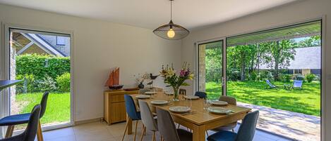 A dining area with a wooden table set for four, surrounded by chairs, with a view of a lush garden and a sailboat model on a cabinet.