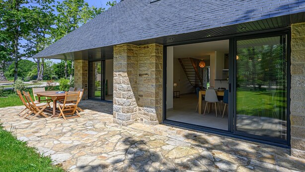 A modern house with a stone exterior, a dining area with a table and chairs, and a large sliding glass door leading to an interior space. - Les Dryades - Villa familiale proche plage (Sarzeau)