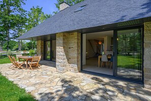 Een modern huis met een gevel van natuursteen, een eetruimte met een tafel en stoelen, en een grote schuifglazen deur die leidt naar een binnenruimte.