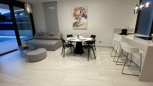Een moderne woonkamer met een bank, een eettafel en stoelen. Er is een keuken met een spoelbak en een aanrecht. Een schilderij hangt aan de muur.