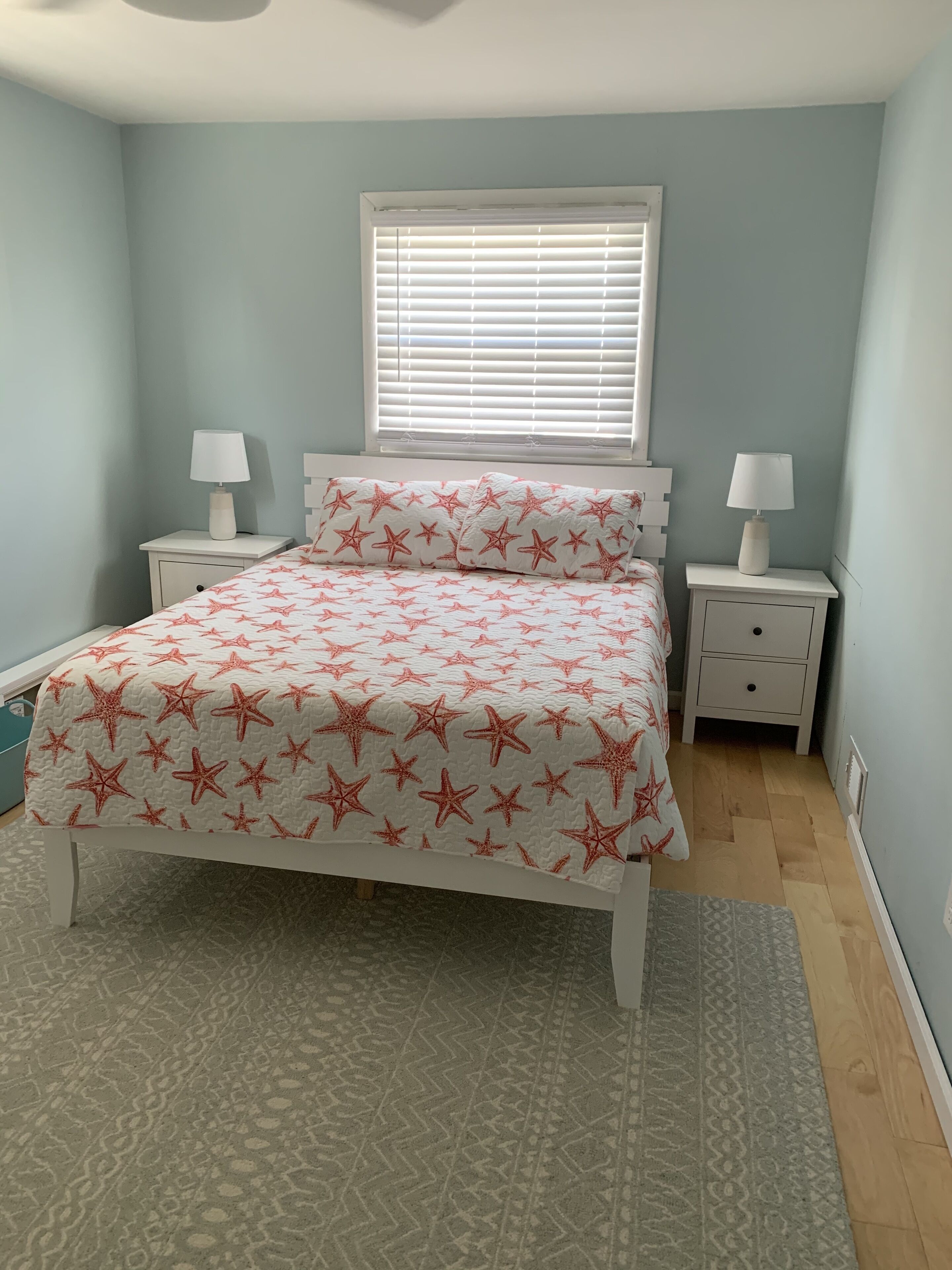 Une chambre à coucher avec un lit, deux tables de chevet avec des lampes, une fenêtre avec des stores et un tapis à motifs.