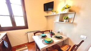 A dining area with a table set for four, chairs with woven seats, a TV mounted on the wall, and a window with blinds. - Family room (Toscana)