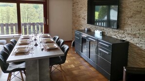 Een eetruimte met een lange tafel voor zes personen, een keuken met een televisie en een uitzicht door een groot raam.